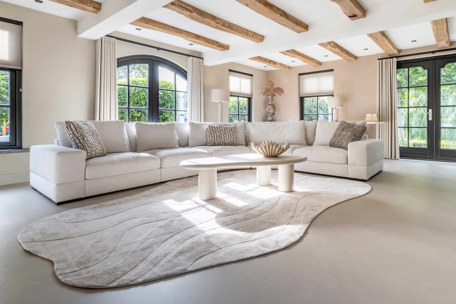 Luxe woonkamer met natuurlijke kleuren, grote ramen en houten plafondbalken voor een warme uitstraling.